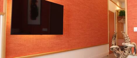A room with a large flat-screen TV mounted on a textured orange wall, a wooden chair with a unique design, and a small table with a candle.