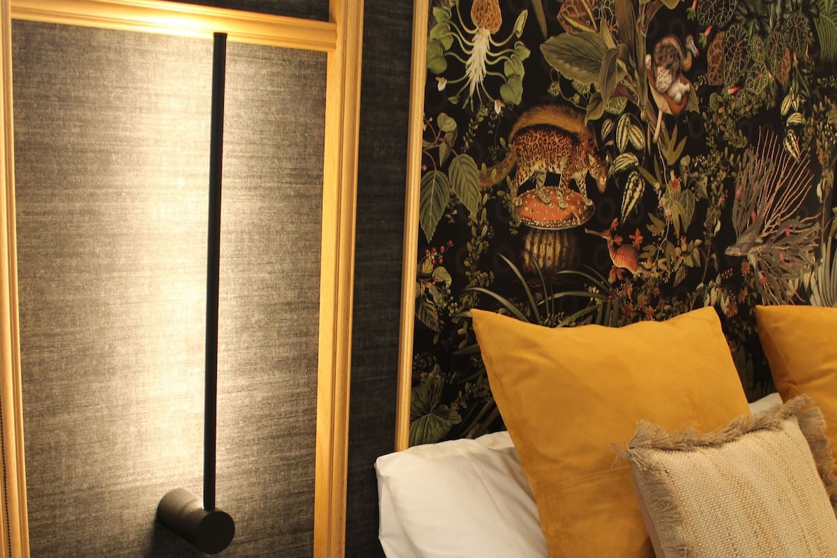 A bedroom with a bed, pillows, a remote control, a small round table, and a wall with a floral tapestry.