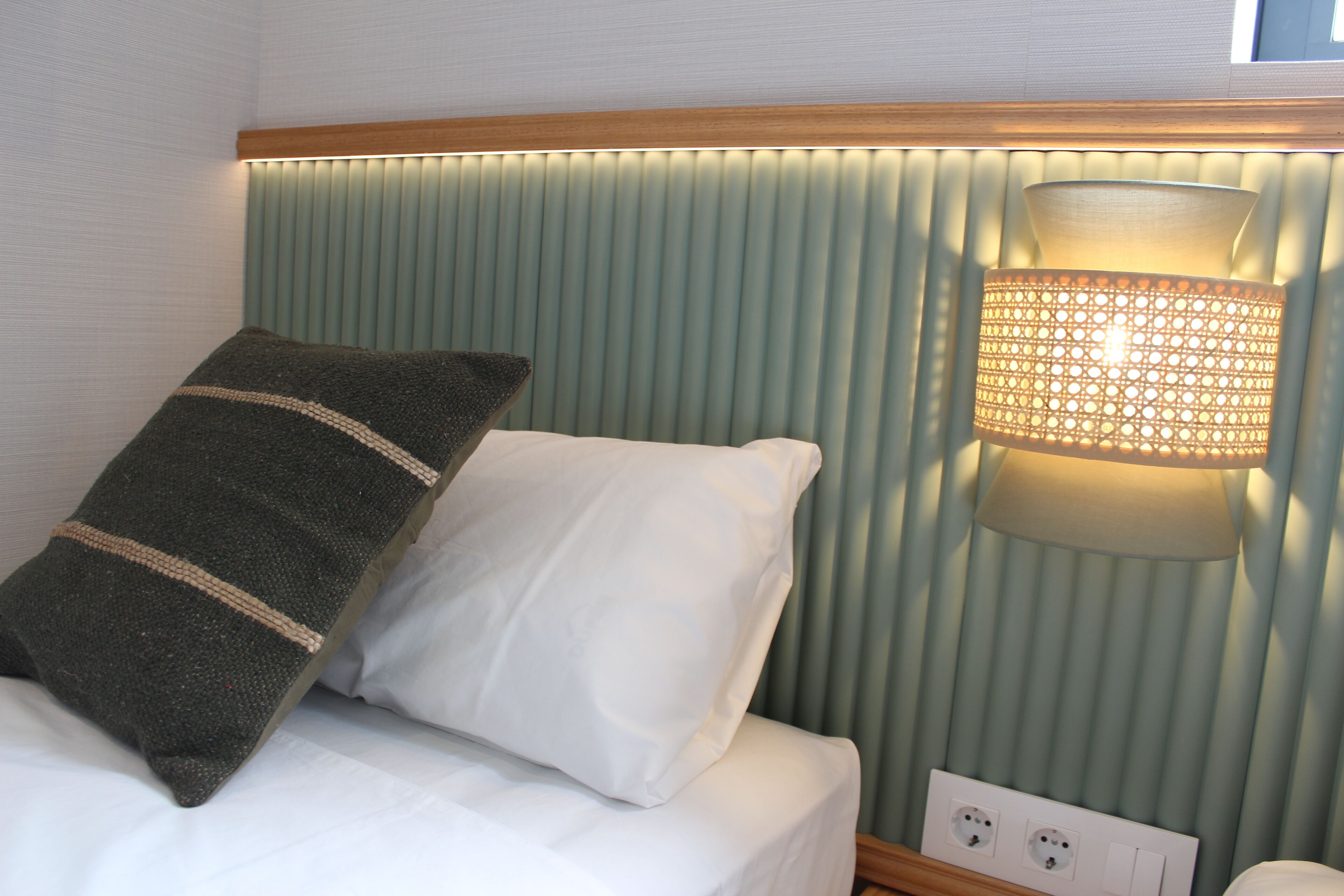 A hotel room with a bed, a nightstand, a wall-mounted lamp, and a power outlet.