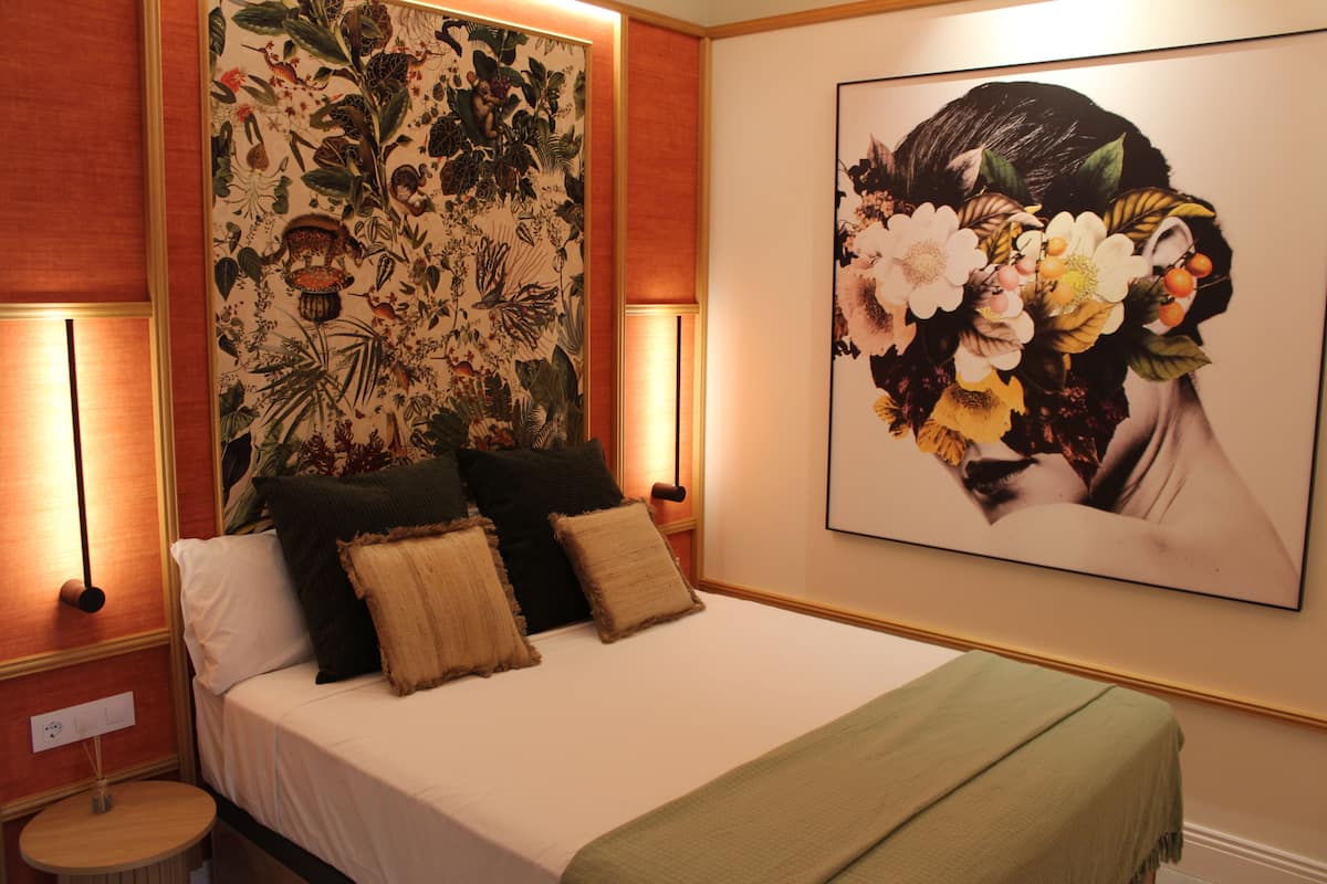 A bedroom with a bed, pillows, a green blanket, a wall tapestry, and two wall-mounted lamps.