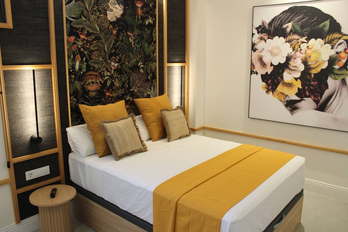 A neatly made bed with a white mattress and a mustard-colored blanket, flanked by two pillows, in a room with a dark wood panel design and a large floral artwork on the wall.