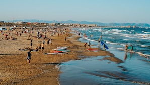 A beach with people walking and standing, and surfboards lined up on the sand. - Promotur Suites - Ayora (Valencia)