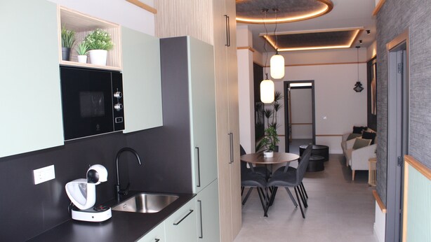 A modern kitchen with a built-in oven, a sink, and a countertop with a food processor. There is a dining area with a table and chairs, and a sofa in the background. - Promotur Suites - Ayora (Valencia)