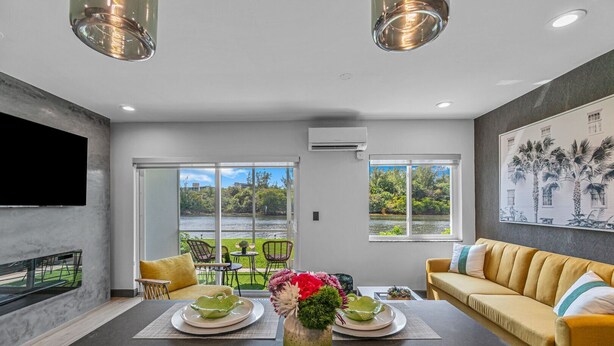 A modern living room with a large window, a flat-screen TV, and a dining area with a table set for two. - 261 #5 Managed By Brampton Park (Deerfield Beach)