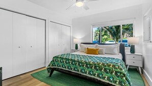 Quarto com cama, mesa de cabeceira, janela com vista para palmeiras e ventilador de teto.