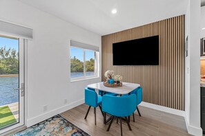 Een moderne eethoek met een witte tafel en blauwe stoelen, een flatscreen televisie bevestigd aan een houten wandpaneel, en uitzicht op een meer door een raam.