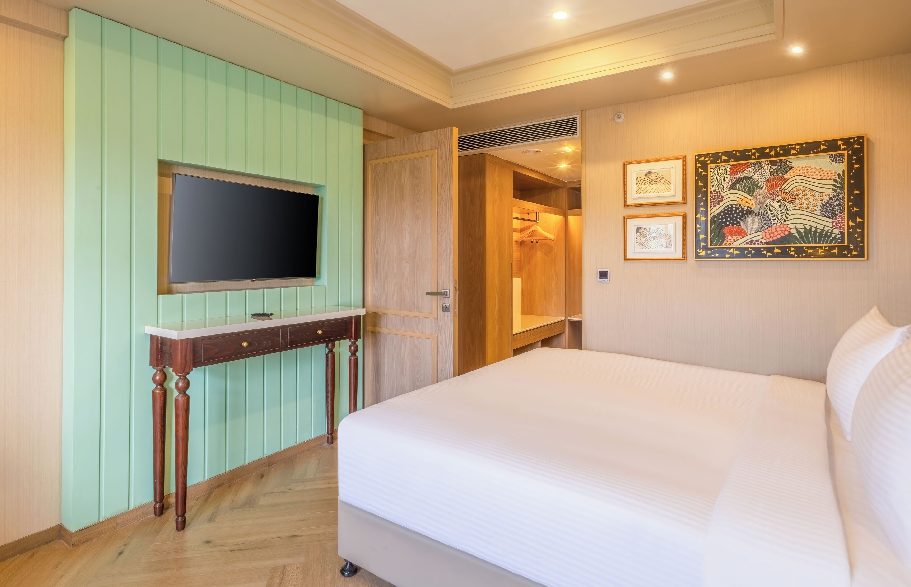 A hotel room with a bed, a television mounted on a wall, a wooden desk, and a wardrobe.