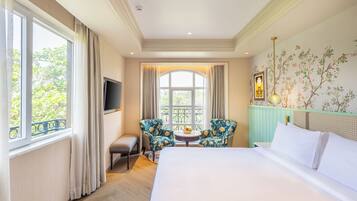 Une chambre d’hôtel avec un grand lit, une télévision, deux fauteuils, une petite table sur laquelle se trouve un saladier de fruits, et une fenêtre avec des rideaux.