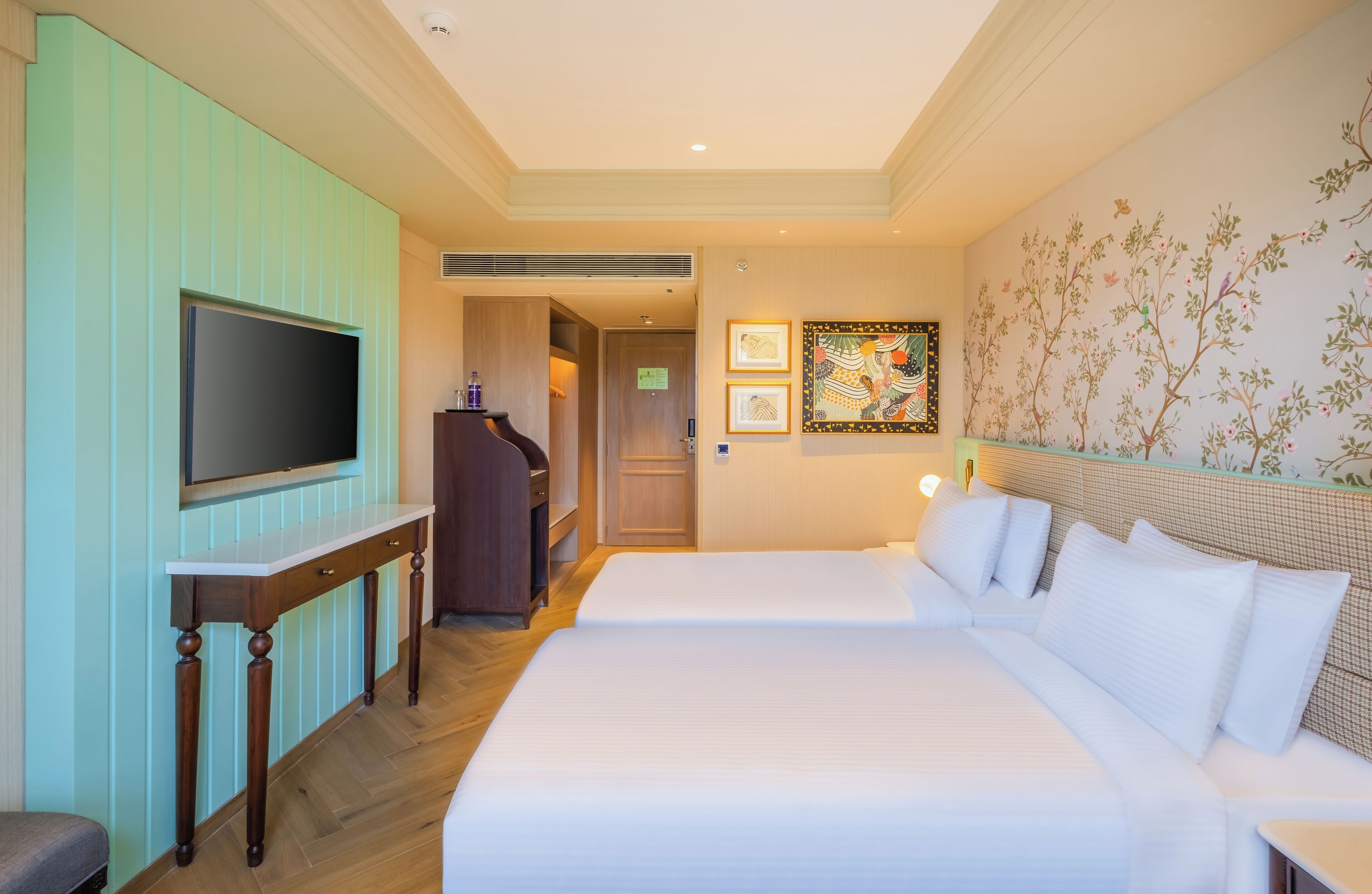 A hotel room with a large bed, a TV on a wooden stand, a desk, and a wall with floral wallpaper.