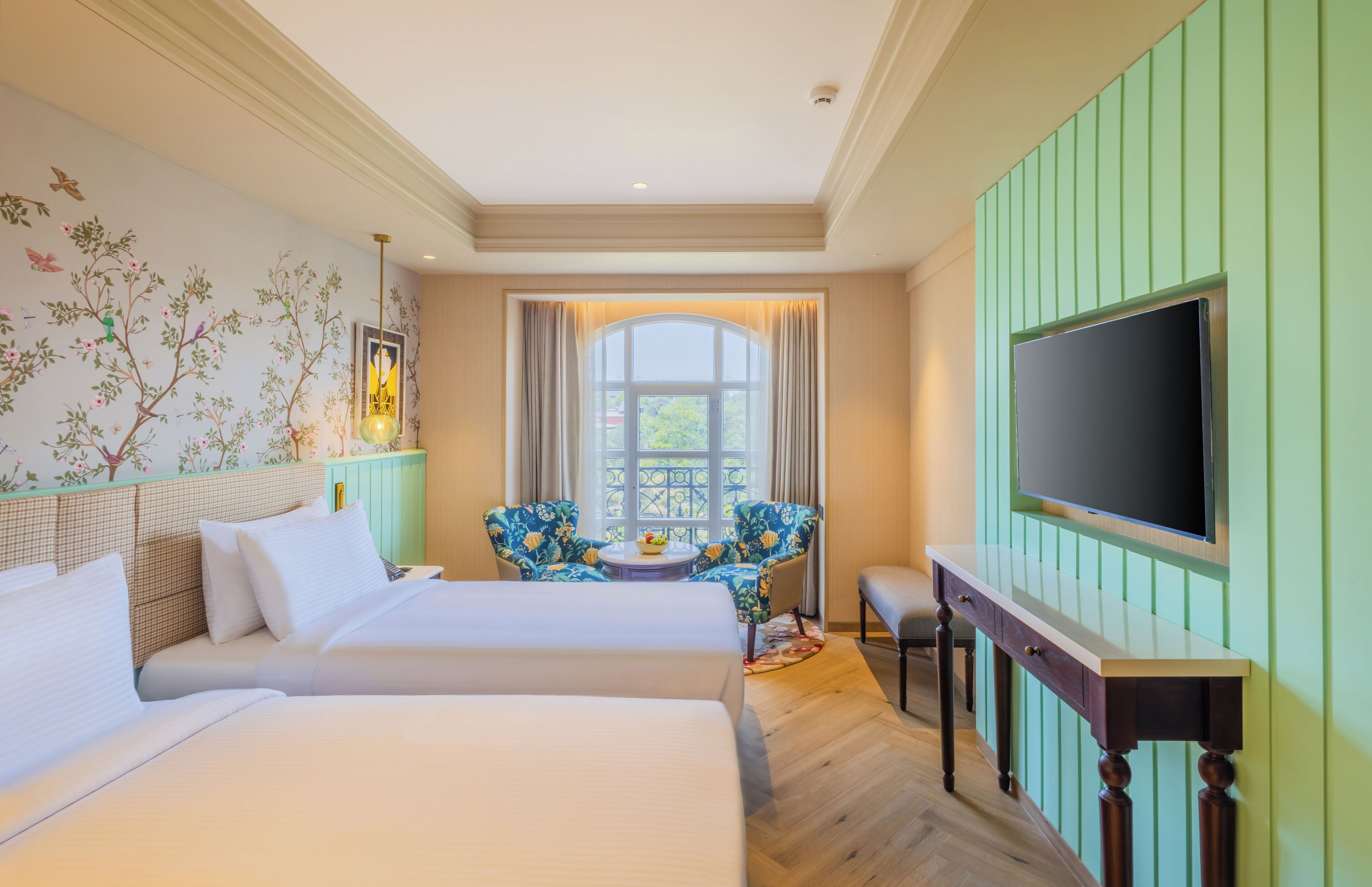 A hotel room with two beds, a TV, a window with curtains, a floral chair, and a small table with a bowl of fruit.