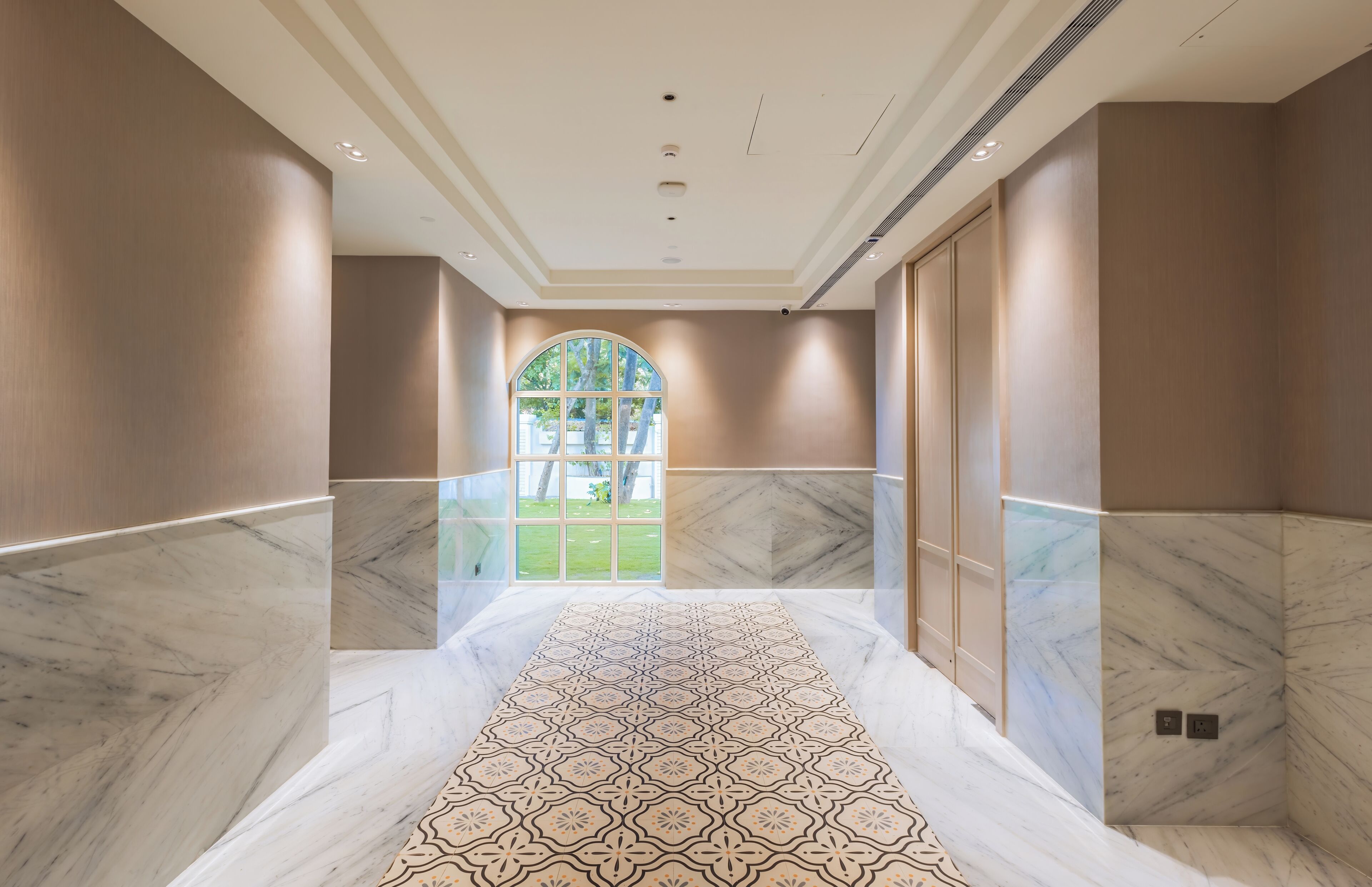 A hallway with marble walls and a patterned floor, leading to a large arched window with a view of a green lawn.