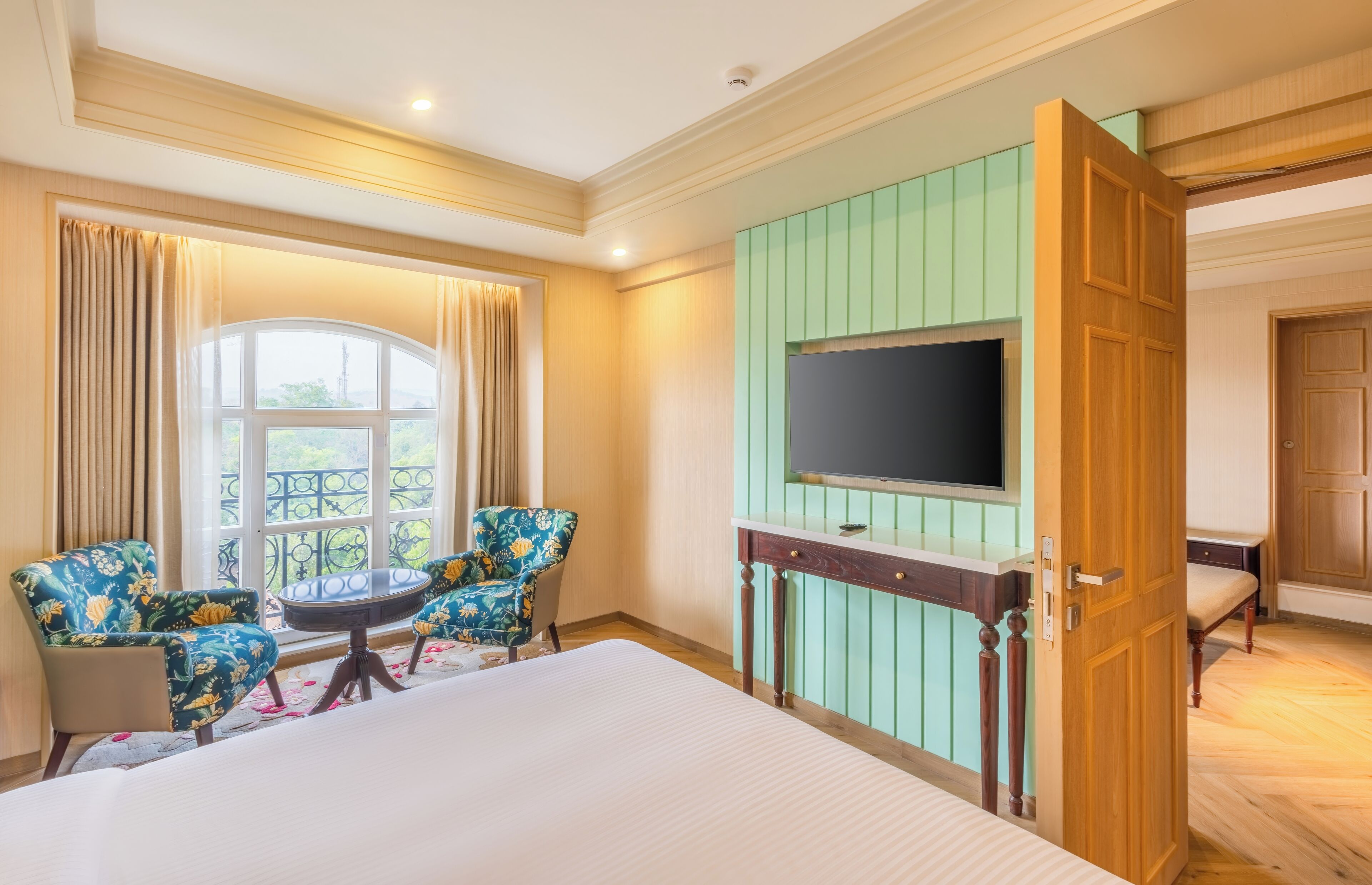 A hotel room with a large bed, a TV mounted on a green wall, two floral armchairs, a small round table, and a window with curtains.