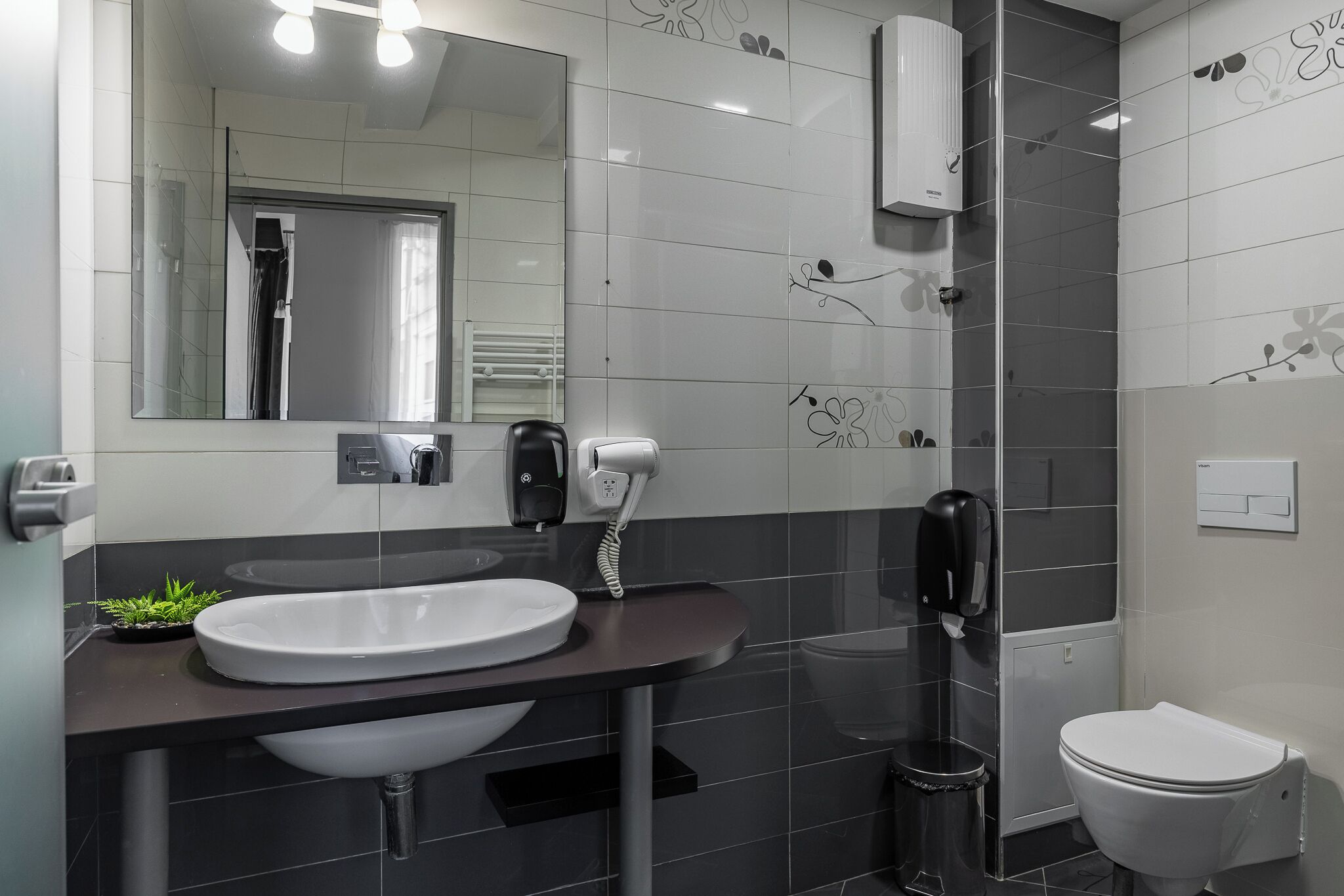 A modern bathroom with a large mirror, a white sink, a black towel dispenser, and a toilet.