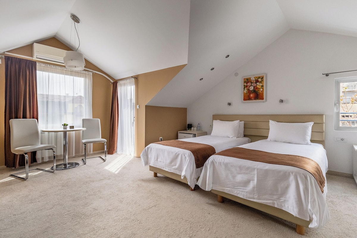 Hotelkamer met twee bedden, een bureau met stoelen, een klein tafeltje en een raam met gordijnen.