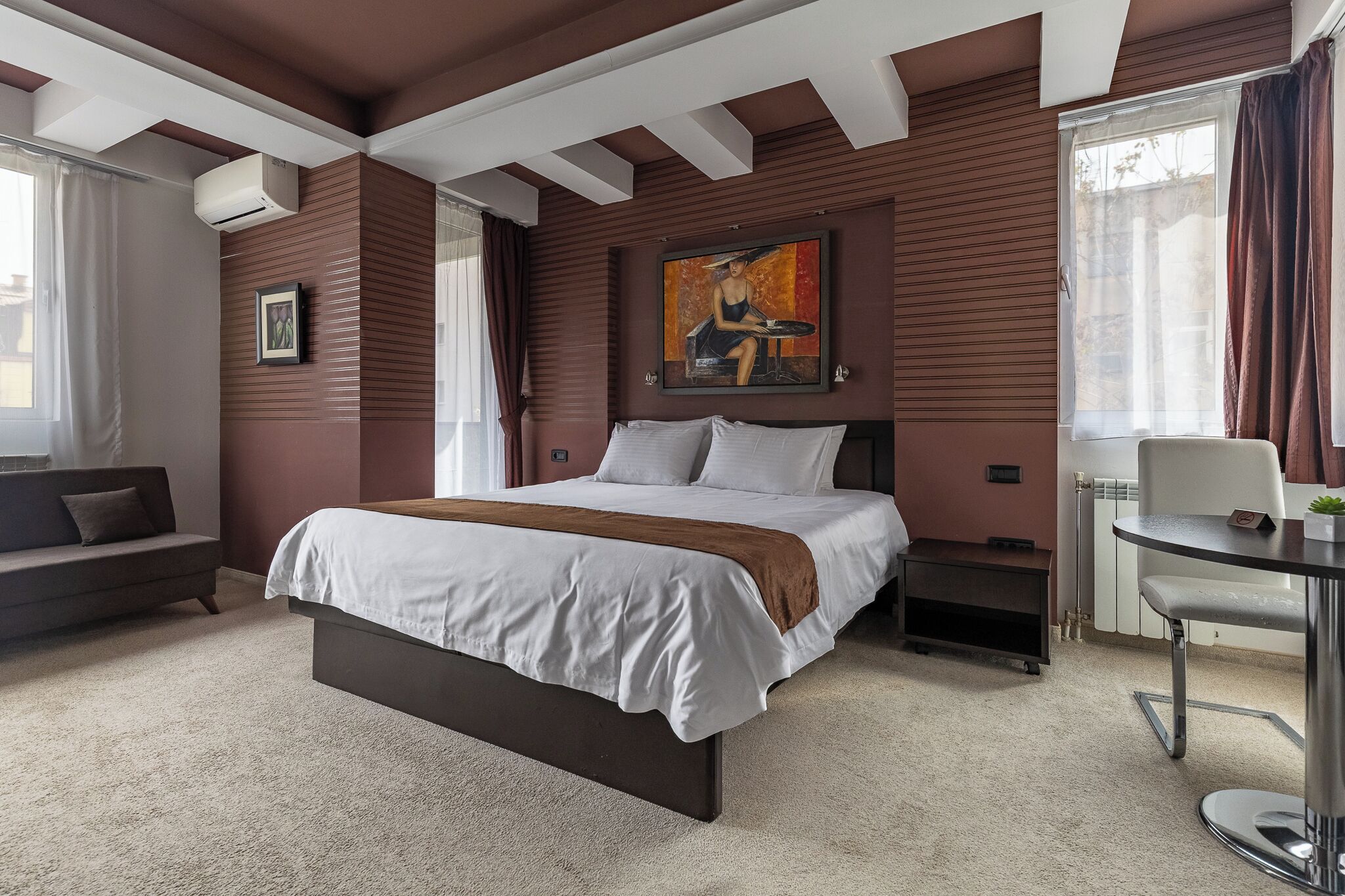 A bedroom with a bed, a sofa, a table, a painting, and a radiator.