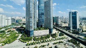 Un ensemble immobilier composé d’immeubles de grande hauteur, dont plusieurs gratte-ciel, entouré de verdure et bordé par une rue très fréquentée où circulent de nombreux véhicules.