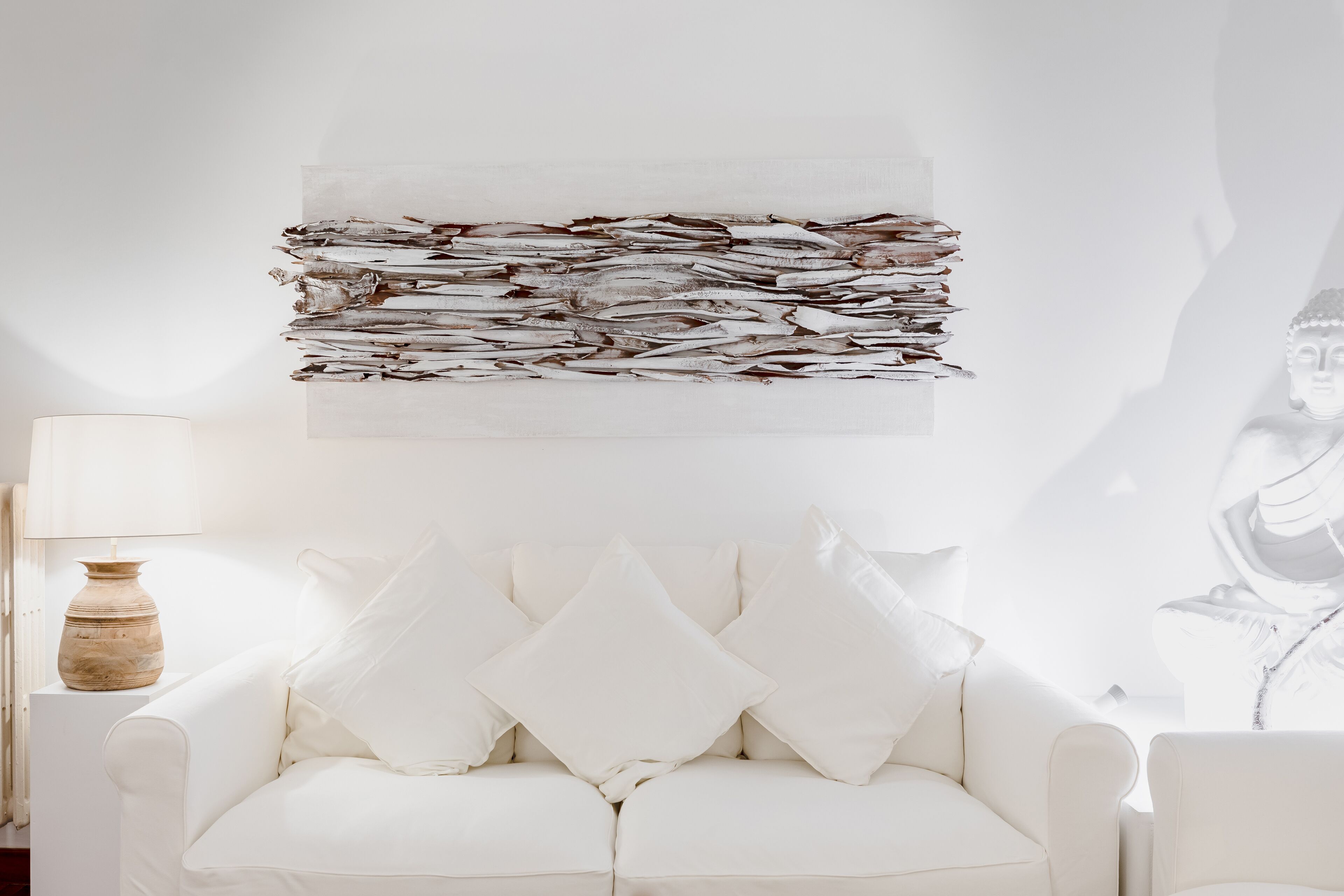 A white sofa with two pillows, a vase, and a wall art piece.