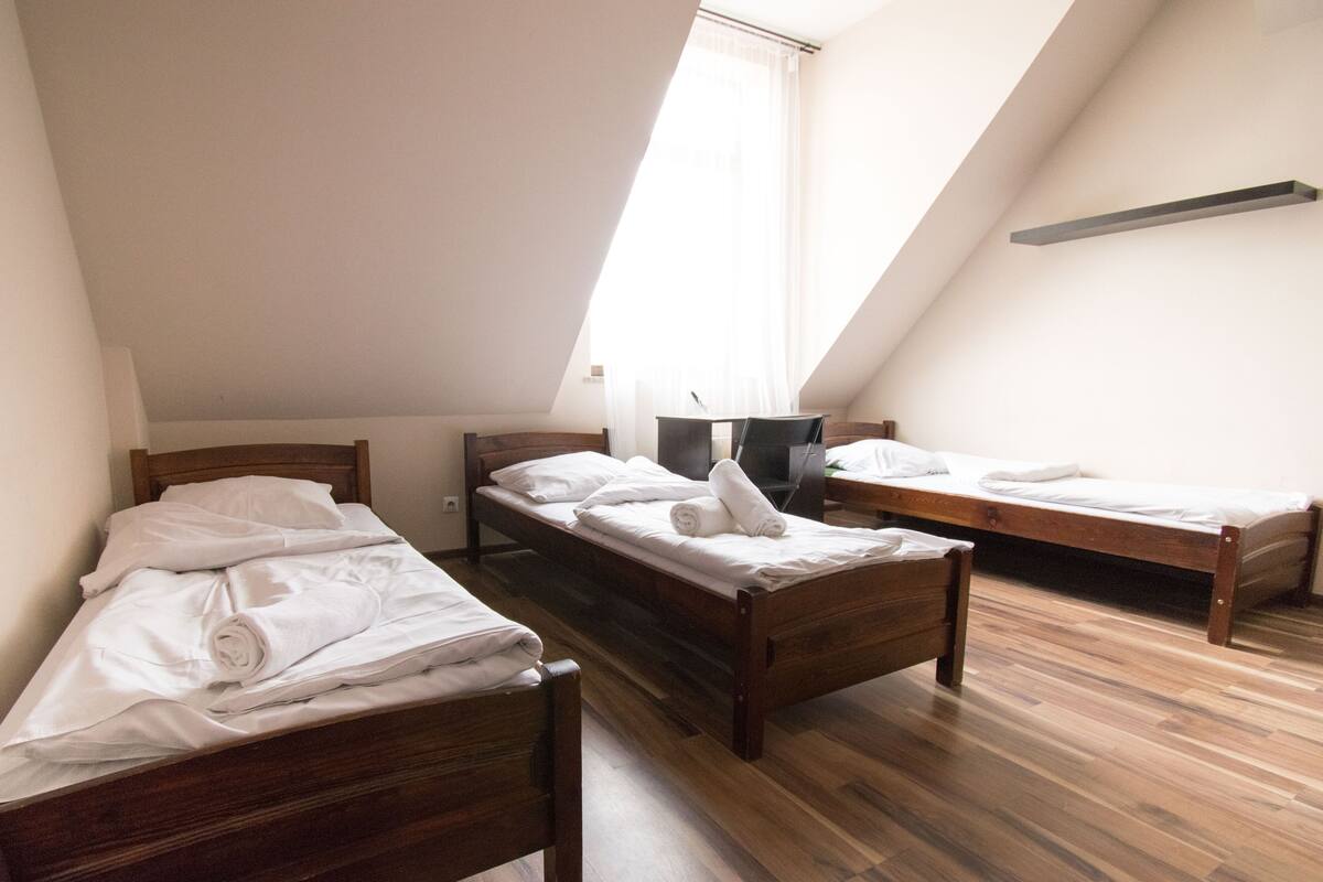 Une chambre de dortoir avec trois lits en bois, un téléviseur posé sur un meuble et une fenêtre ornée de rideaux légers.