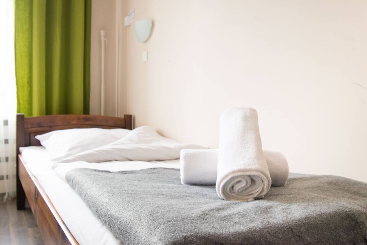 Un lit bien fait, avec une couverture grise et une serviette blanche enroulée, une tête de lit en bois et un rideau vert.