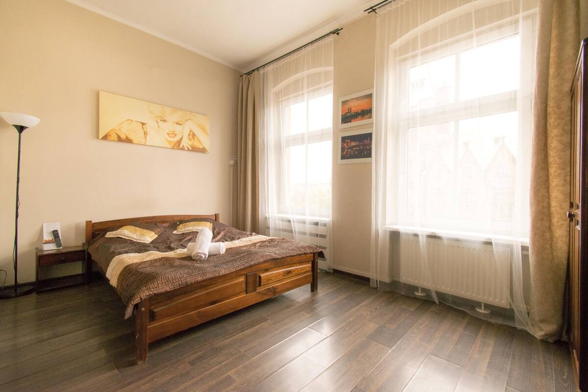 Une chambre à coucher avec un lit en bois, une table de chevet, une lampe et une fenêtre ornée de rideaux légers.