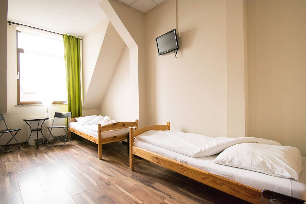 Une chambre avec deux lits en bois, une petite table, des chaises, un téléviseur fixé au mur et une fenêtre avec des rideaux.