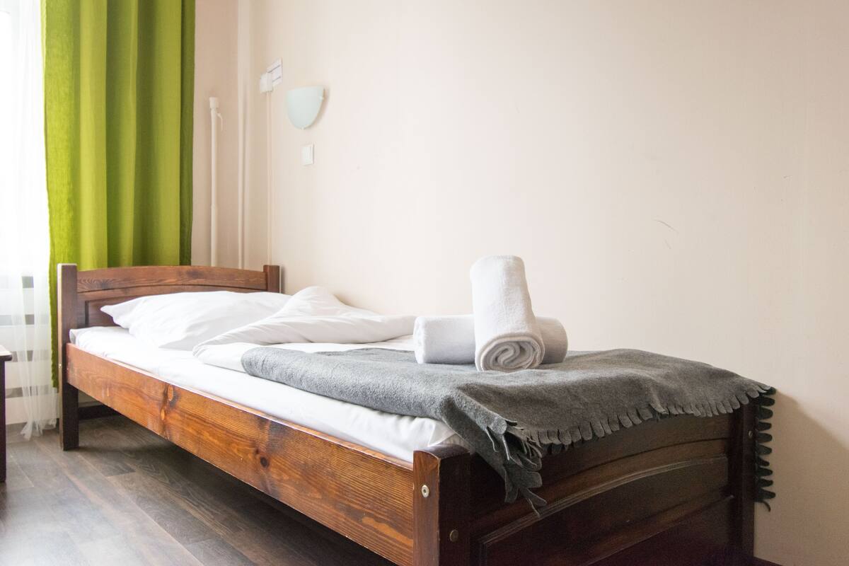 Un lit bien fait, avec des draps blancs et une serviette enroulée, une tête de lit en bois et un rideau vert, dont on aperçoit une partie.