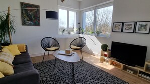 Ein modernes Wohnzimmer mit einer Couch, zwei Sesseln, einem Couchtisch, einem Fernseher und einem großen Fenster mit Pflanzen.