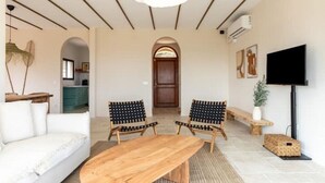 Un salon moderne avec une table à manger en bois, deux chaises à rayures noires et blanches, un canapé blanc, un banc en bois, un téléviseur fixé au mur et une porte en arc donnant sur une autre pièce.