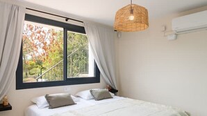 Une chambre à coucher avec un lit, des rideaux blancs, une suspension en osier et une fenêtre donnant sur des arbres.