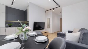 Un comedor moderno con una mesa negra dispuesta para cuatro personas, un televisor de pantalla plana y un sofá gris al fondo.