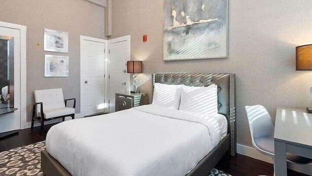 A hotel room with a large bed, a bedside table with a lamp, a chair, and a painting on the wall. - Bay VIllage Suite, Ready To Host! (Boston)