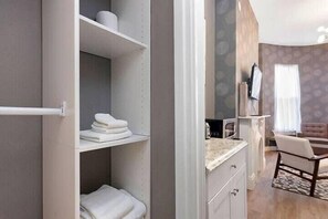 Habitación de hotel con armario empotrado, kitchenette pequeña, zona de estar con una silla y un ventanal grande con cortinas.