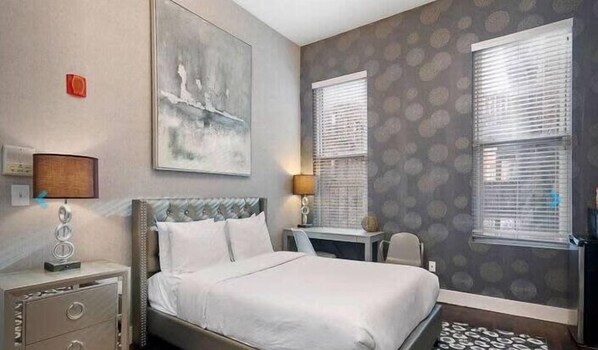 A hotel room with a bed, a desk, two lamps, and a large abstract painting on the wall. - Studio Awaits Your Next Stay! (Boston)