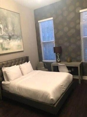 A hotel room with a bed, a desk, and two windows with blinds. - Studio Awaits Your Next Stay! (Boston)