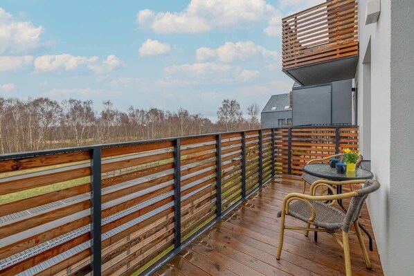 Una terraza de madera con una mesa y sillas, una cerca metálica y un edificio con techo inclinado al fondo.