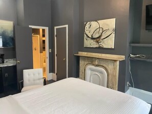A bedroom with a bed, a white chair, a fireplace, and a painting on the wall. - Affordable & Simple Chic Studio (Boston)