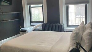 A hotel room with a bed, two windows with blinds, a radiator, a flat-screen TV mounted on the wall, and a leopard print carpet.