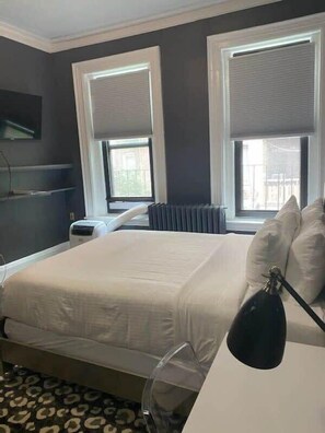 A hotel room with a bed, two windows with blinds, a radiator, a desk with a lamp, and a patterned carpet. - Affordable & Simple Chic Studio (Boston)