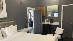 A hotel room with a bed, a desk, a chair, a kitchenette, and a door leading to another room.