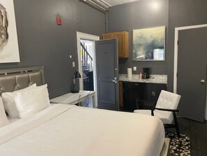 A hotel room with a bed, a chair, a desk, a kitchenette, and a door leading to another room. - Affordable & Simple Chic Studio (Boston)
