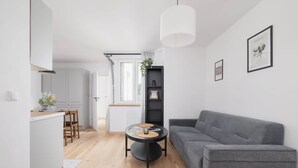 Un salon moderne avec un canapé gris, une table basse ronde et un coin cuisine doté de meubles blancs et d’un plan de travail en bois.