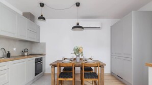 Une cuisine compacte avec des meubles blancs, une table à manger en bois et des chaises, ainsi qu’un luminaire suspendu noir.
