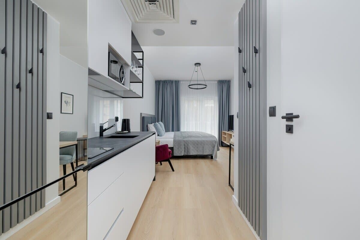 A modern, compact hotel room with a kitchenette, a bed with a gray headboard, and a large window with curtains.