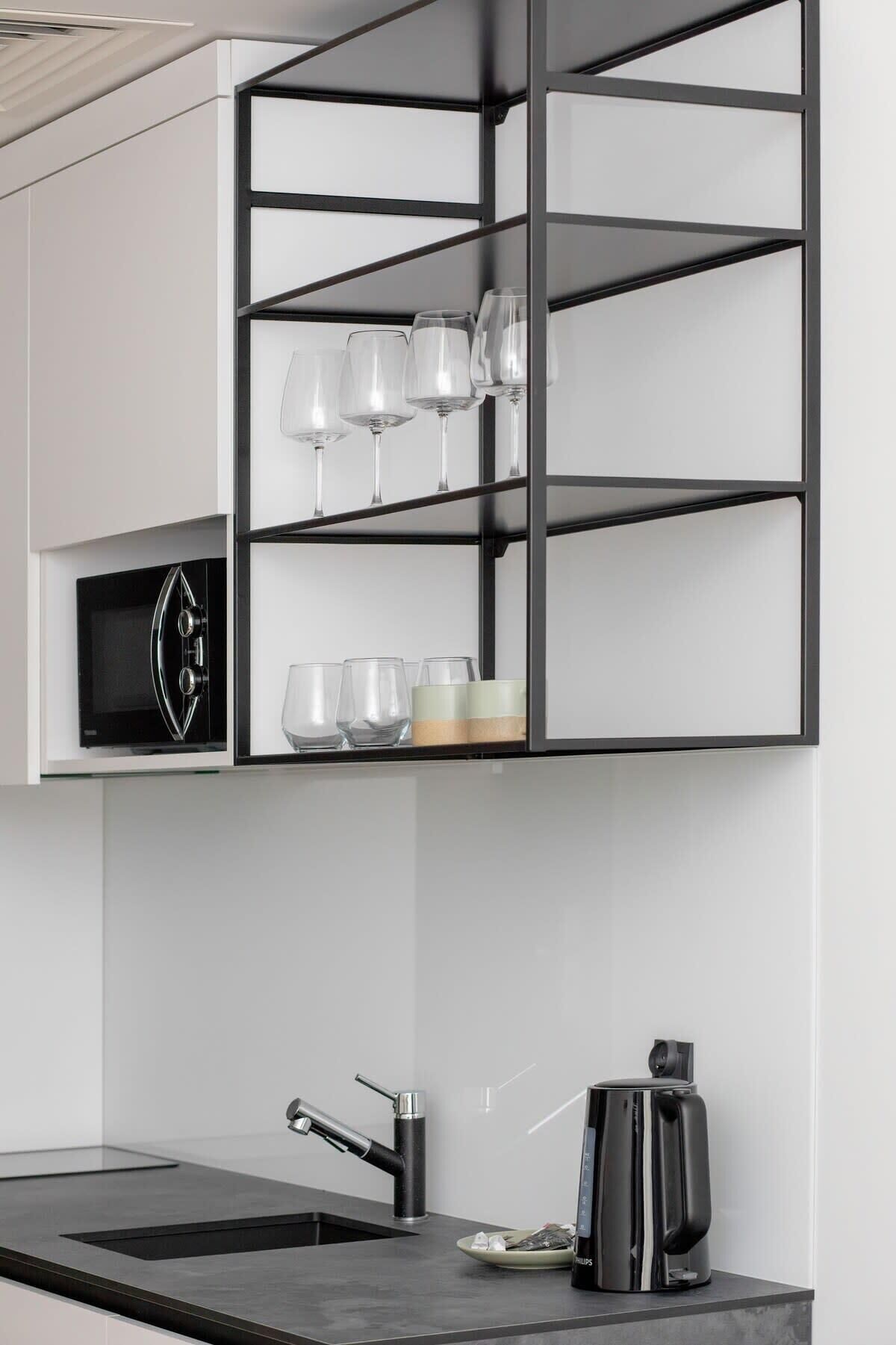 A modern kitchen with a built-in microwave, a coffee maker, and a glass shelf with wine glasses.