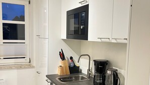A modern kitchen with white cabinets, a black countertop, a built-in microwave, a coffee maker, and a window with a view of the city. - Design Apartment in Carolinensiel (Wittmund)
