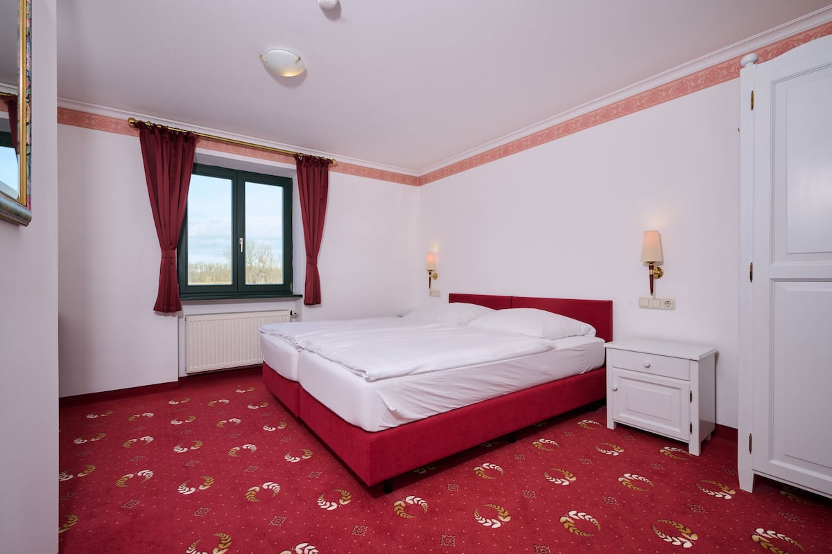 Une chambre avec un lit rouge, des murs blancs, une fenêtre avec des rideaux rouges, une table de chevet blanche et une porte blanche.