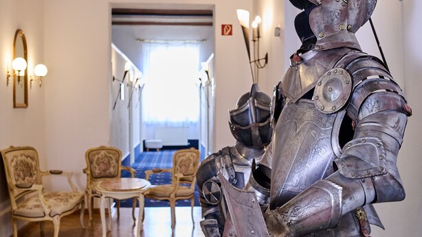 A room with a medieval knight statue, wooden chairs, a blue carpet, and a chandelier. - Tafernwirtschaft Hotel Schönbrunn (Landshut)