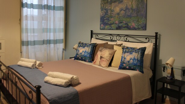 A neatly made bed with a blue and beige bedspread, a painting of a landscape on the wall, and a small bedside table with a lamp.
