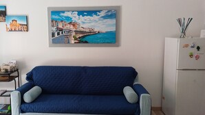 A living room with a blue sofa, a small table with books and a lamp, and a refrigerator with magnets.