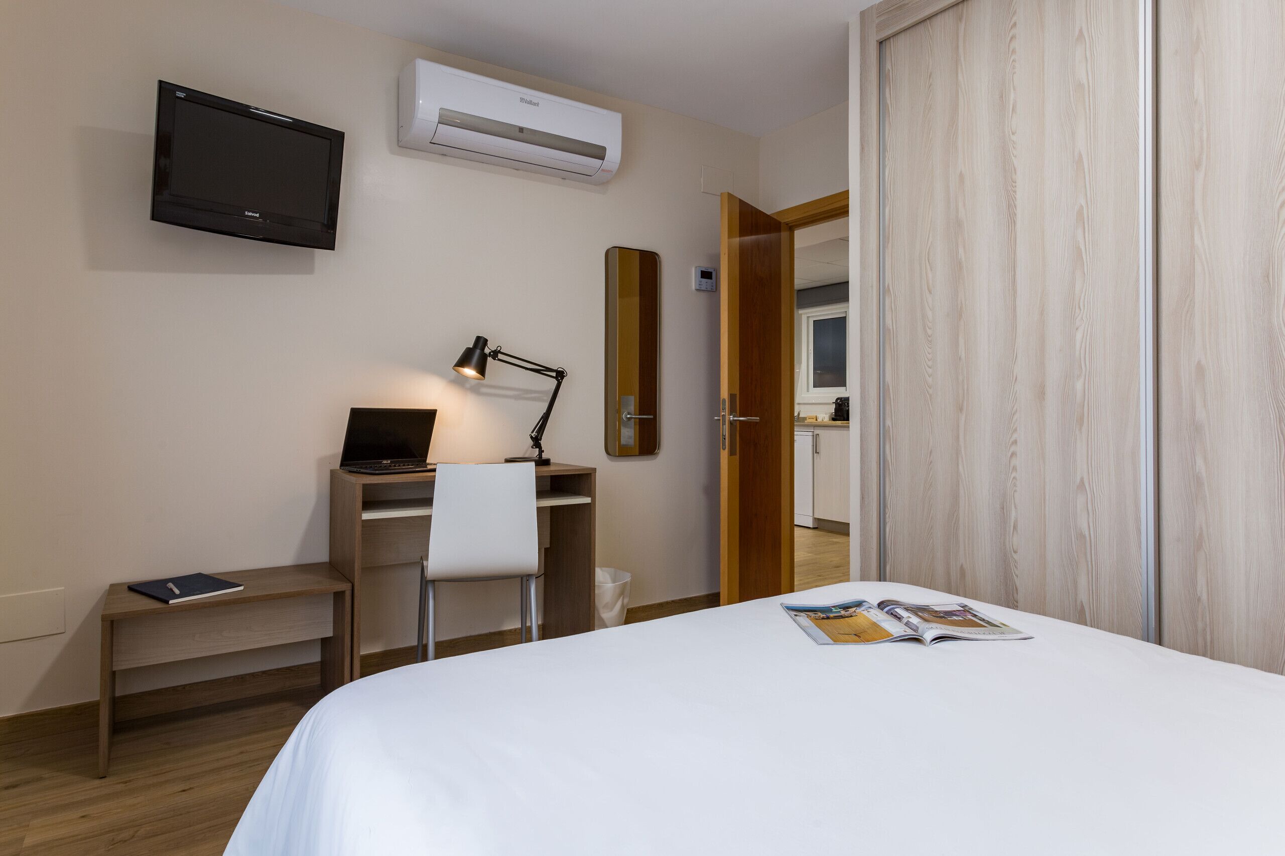 Une chambre d’hôtel comprenant un lit, un bureau avec un ordinateur portable, une chaise, une lampe de bureau, un téléviseur à écran plat, un climatiseur et une armoire en bois.